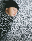 A baby in a beanie swaddled, laying on top of the quilt