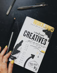 Artistic shot of a hand touching the creative planner on a desk with pens around it.