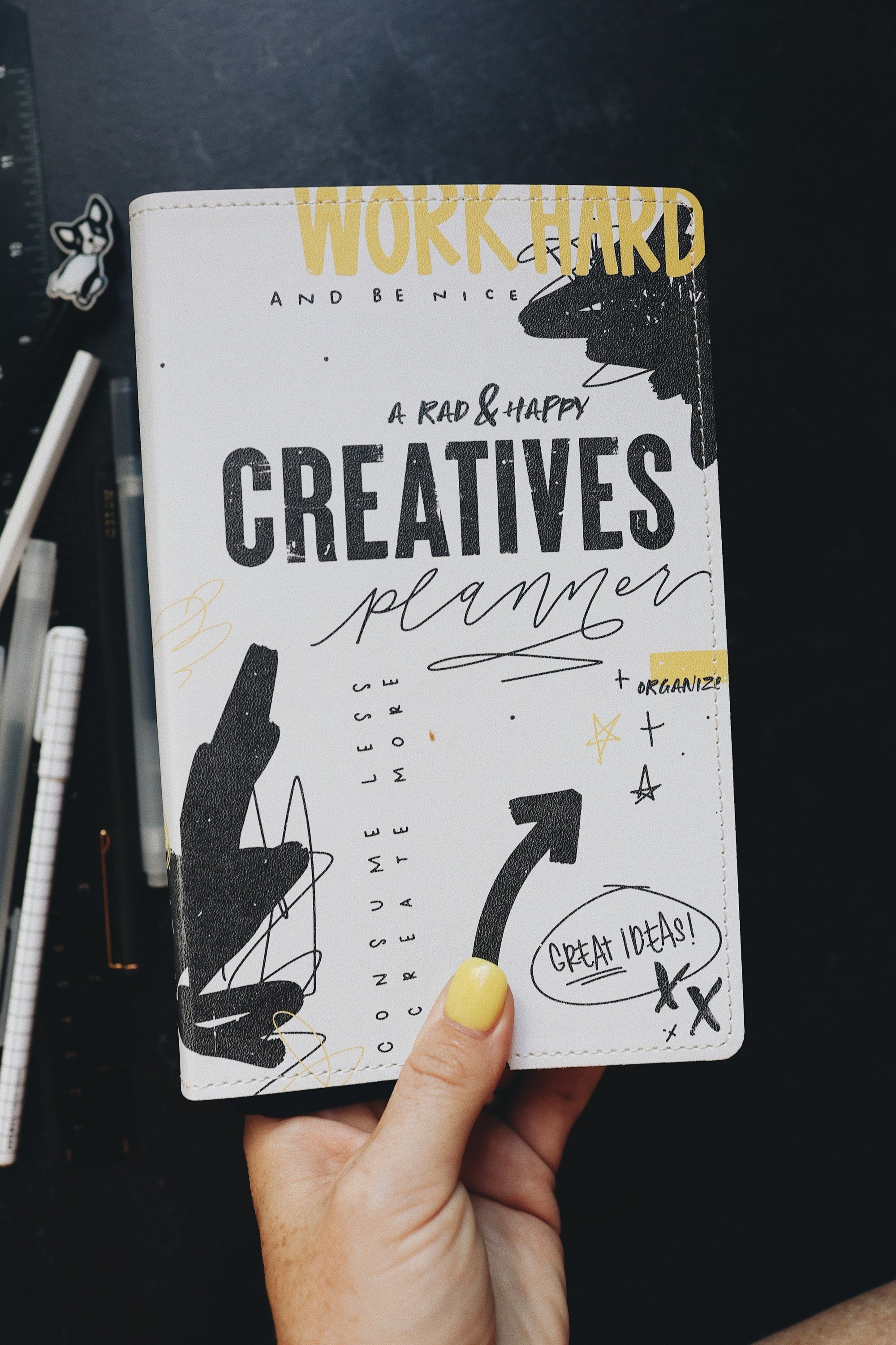 A hand holding the creative planner to show the cover. Desk top in background with pens and a ruler.