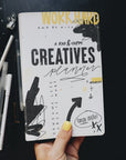 A hand holding the creative planner to show the cover. Desk top in background with pens and a ruler.