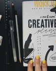 Artistic shot of the creative planner next to a ruler and pens, with three fingers on the bottom of the cover.