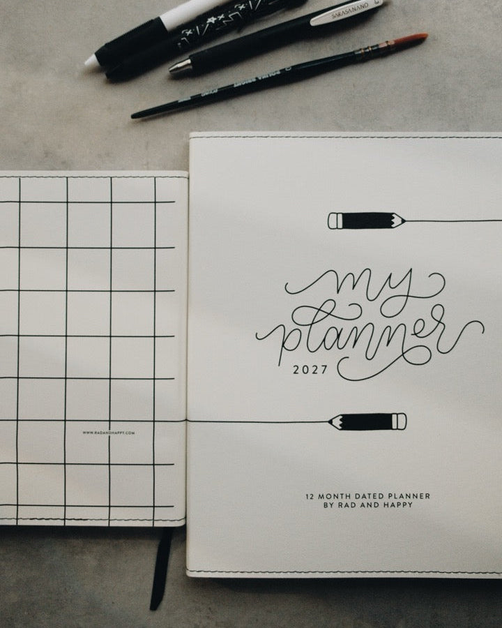 A white 12-month grid planner cover with 'my planner 2027' in black script, shown open on a gray surface alongside pens, a pencil, scissors, and a black ribbon bookmark.