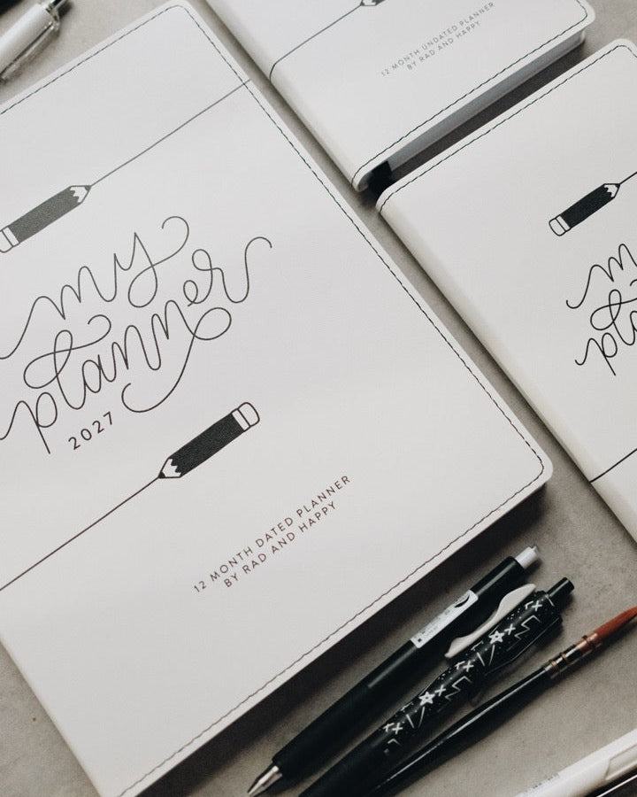 Three white leather-bound planners with black script lettering reading 'my planner 2027' are arranged on a light gray surface with black and white pens around them.