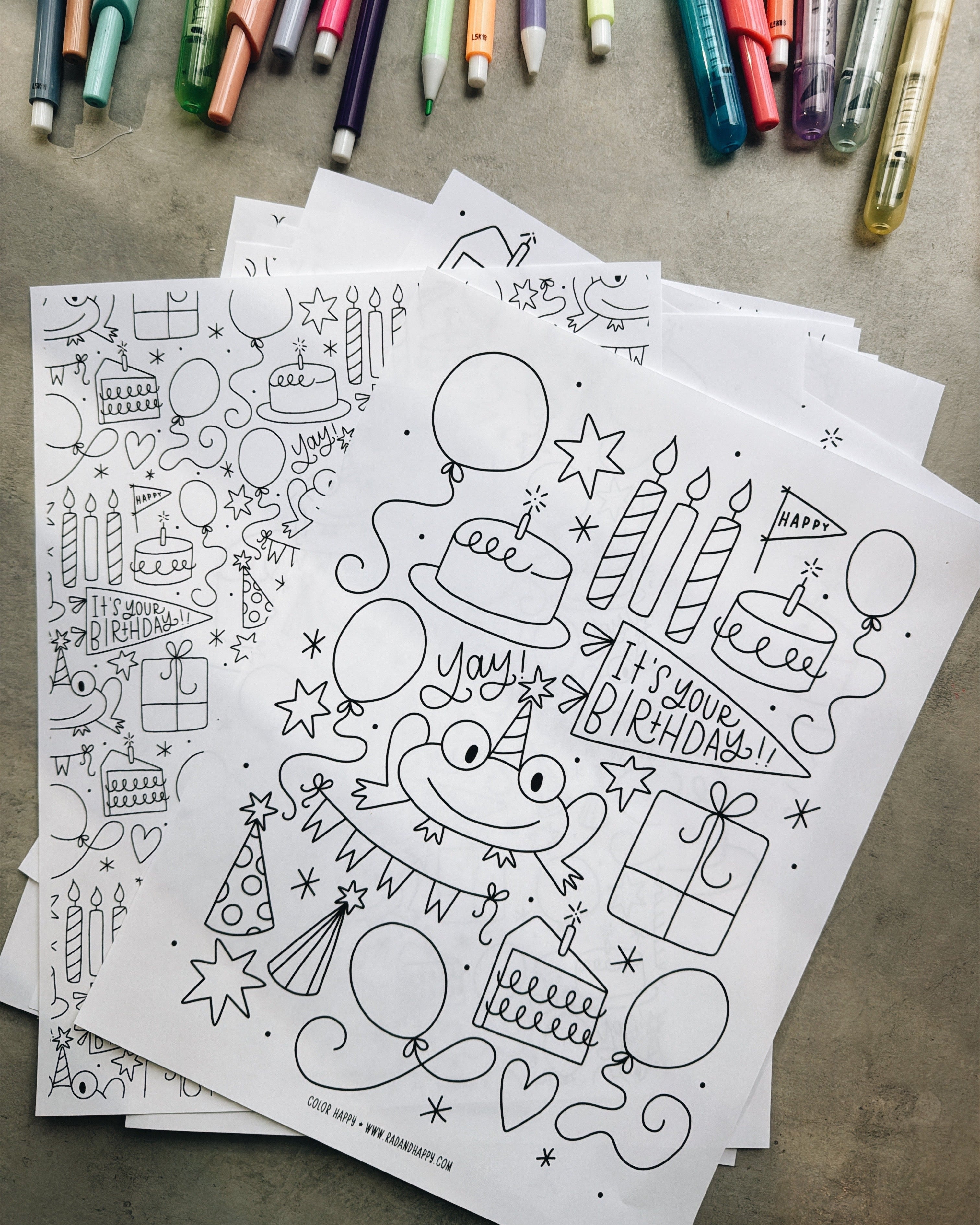 Coloring sheets with birthday-themed illustrations on a textured surface with markers in the background.