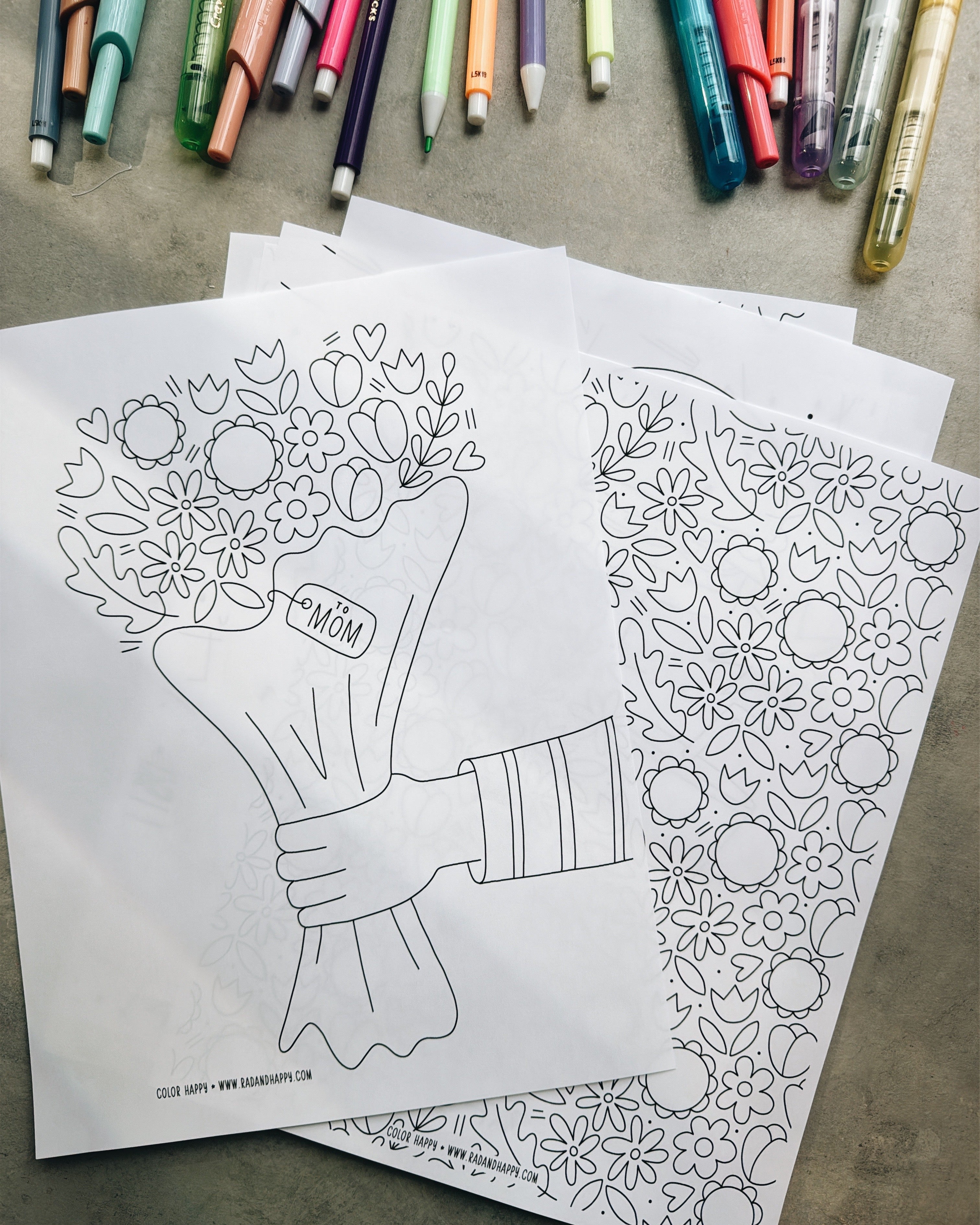 Two coloring pages with floral designs on a textured surface with markers in the background.