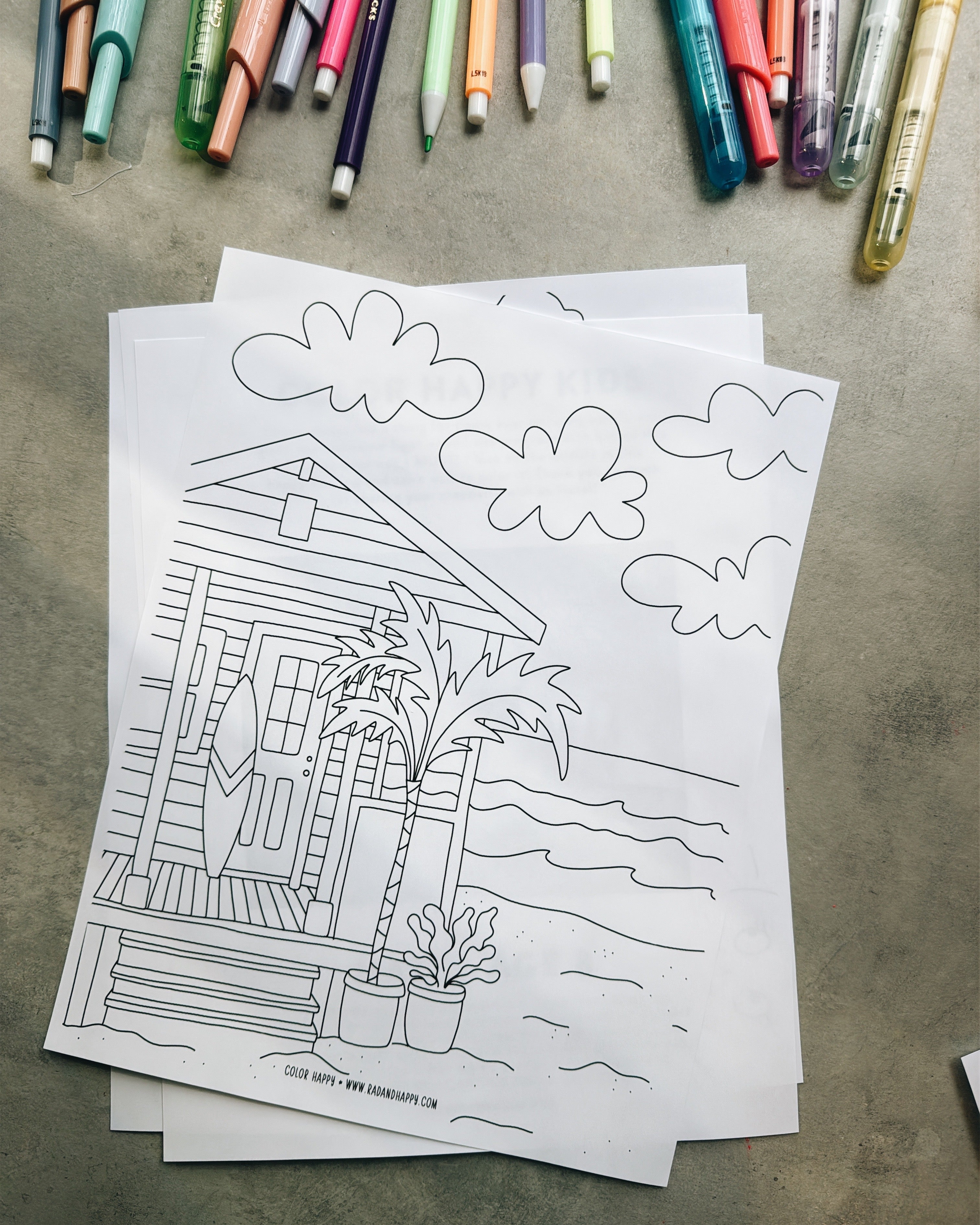 Coloring page of a beach hut with palm trees and clouds, surrounded by markers on a concrete surface.
