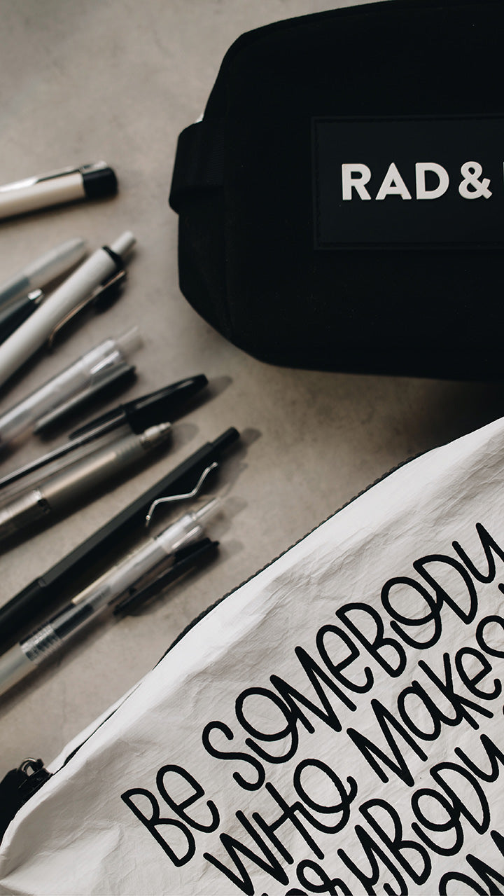 Artistic shot of the white tyvek zipper pouch in the lower part of the photo with the black rad & happy zipper pouch in the upper part sitting on a gray surface surrounded by pens