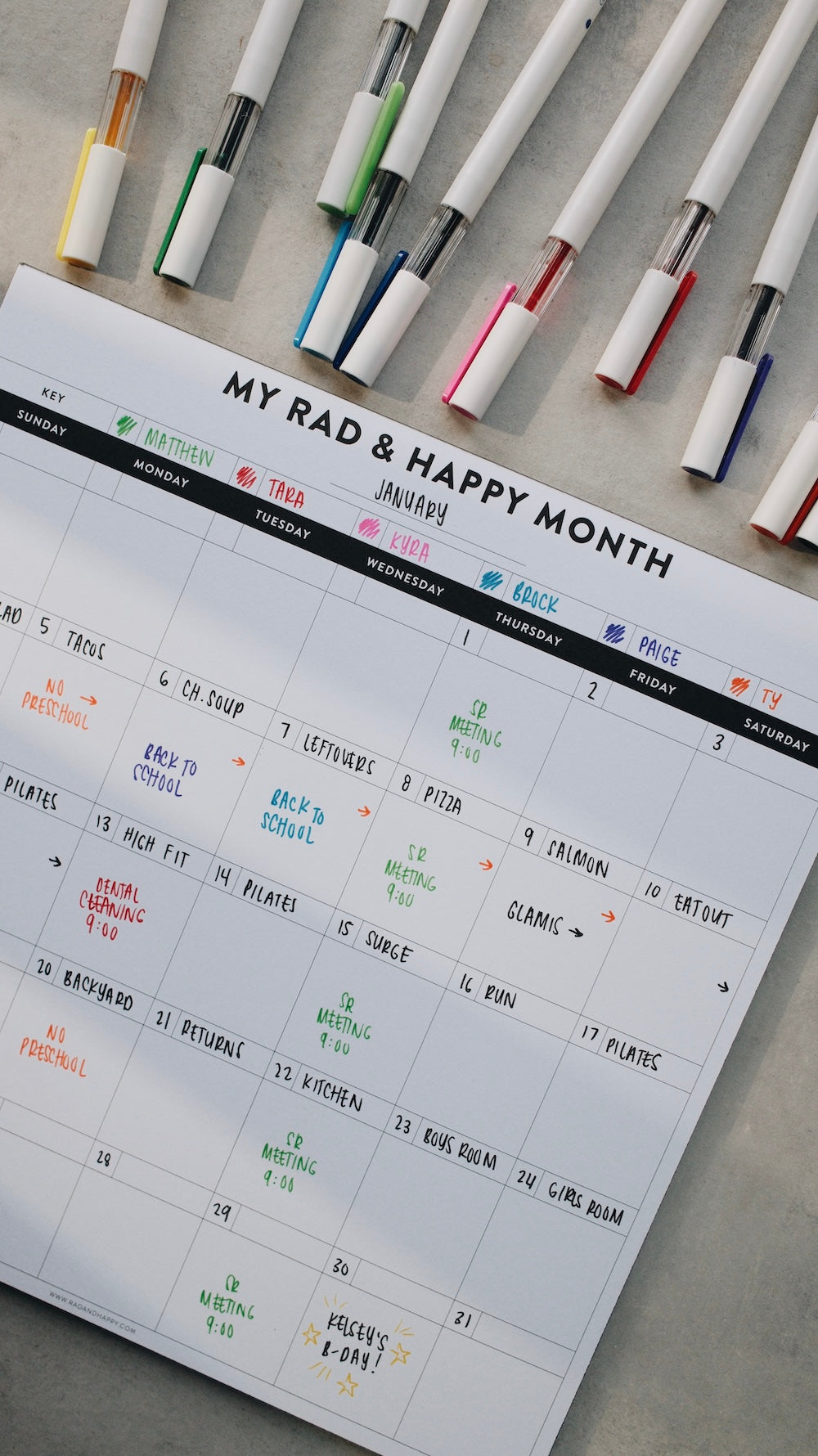 Month notepad with 'My Rad & Happy Month' text, surrounded by colorful markers on a gray surface.