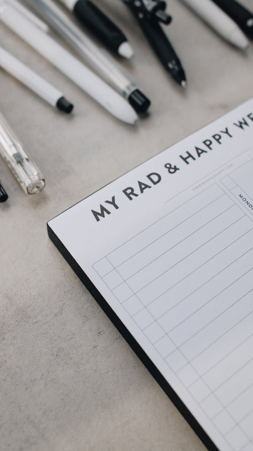 Artistic shot of the corner of a notepad with 'My Rad & Happy Writing' text and various pens on a light surface