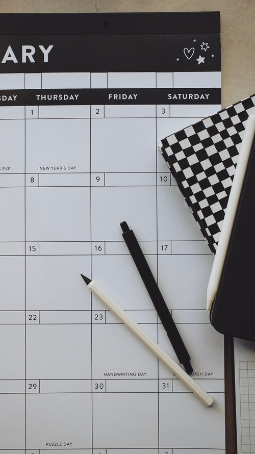 Pens and planners laying on top of a calendar on a light surface