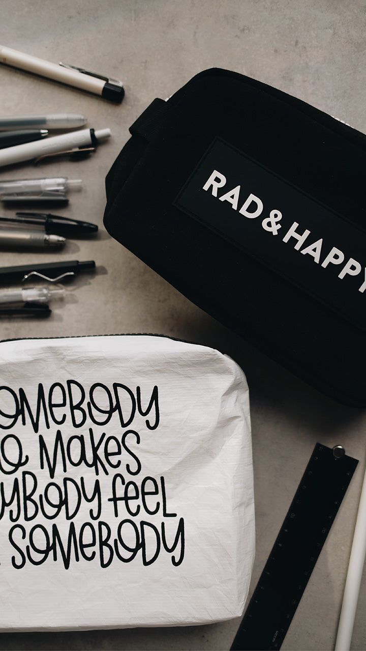 Artistic shot of the white tyvek zipper pouch in the lower part of the photo with the black rad & happy zipper pouch in the upper part sitting on a gray surface surrounded by pens