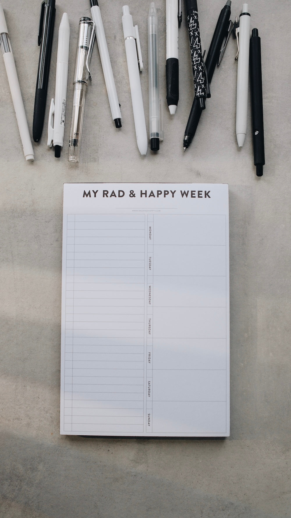 Notepad titled 'My Rad & Happy Week' with pens on a concrete surface