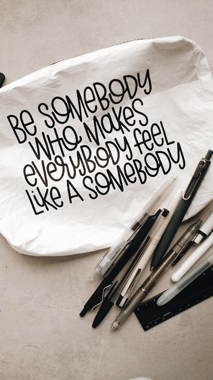White tyvek zipper pouch on a gray surface with pens and the text Be Somebody who makes everybody feel like a somebody in black text