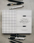 A white grid planner cover with the words 'my planner' in black script, shown open on a gray surface with several pens and pencils arranged around it.