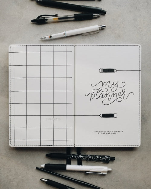 A white grid planner cover with the words 'my planner' in black script, shown open on a gray surface with several pens and pencils arranged around it.
