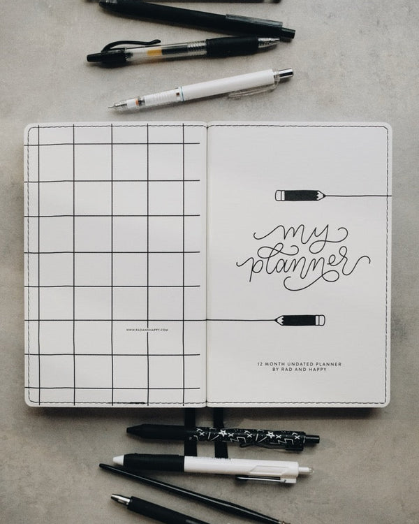 A white grid planner cover with the words 'my planner' in black script, shown open on a gray surface with several pens and pencils arranged around it.