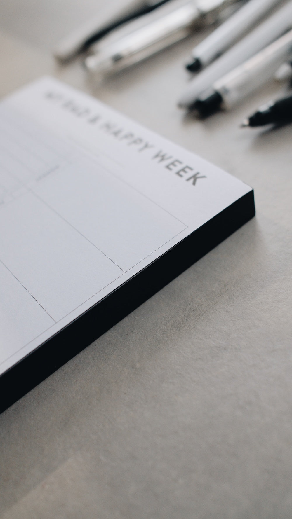 Artistic shot of the corner of a notepad with 'Happy Week' text on a light gray surface with pens in the background