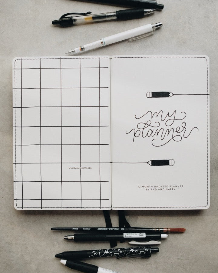 An open white planner with a black grid on the left page and 'my planner' written in script on the right page, surrounded by several pens on a light gray textured surface.