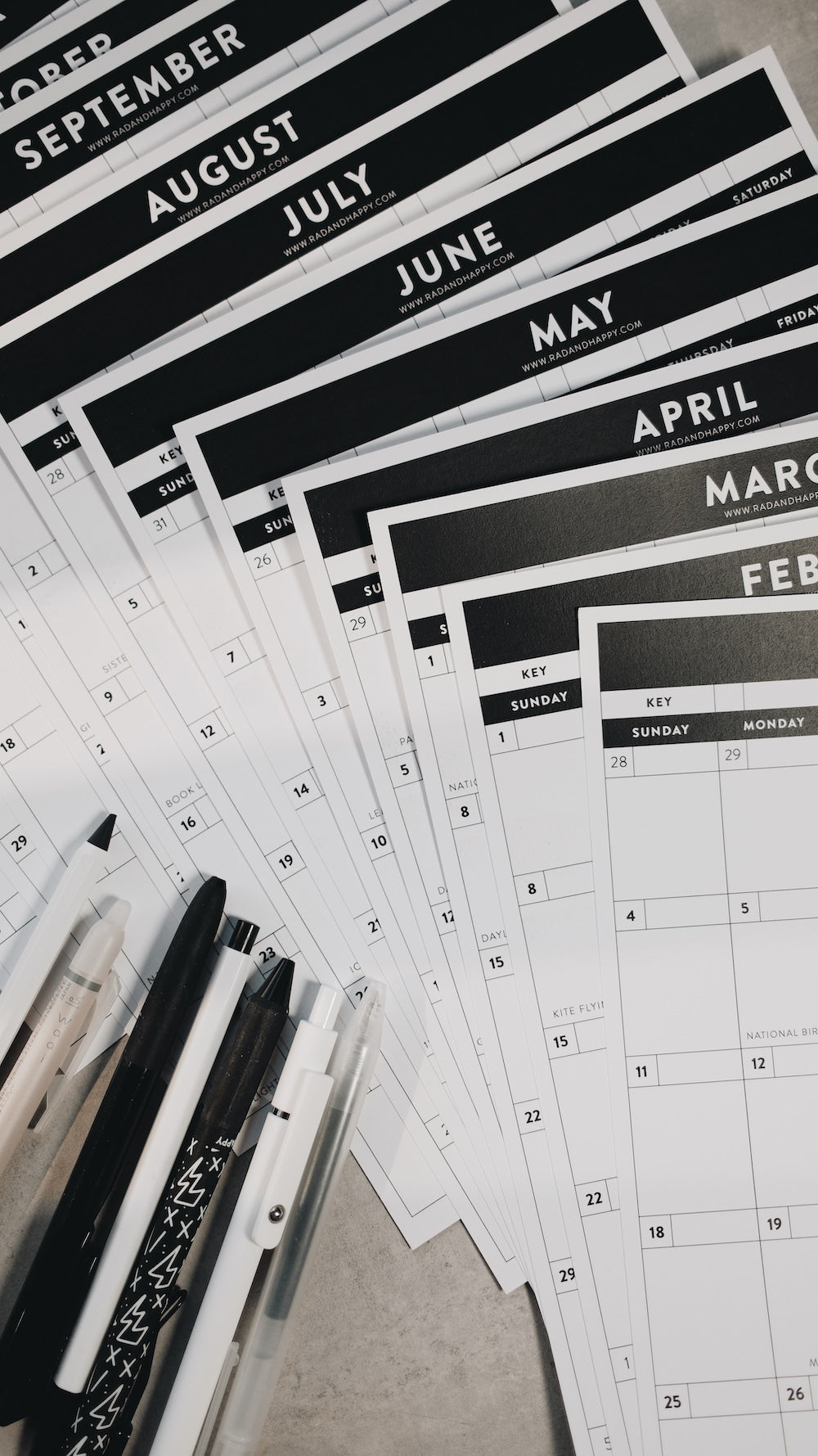 Calendar sheets with months printed on them, arranged on a flat surface with pens.