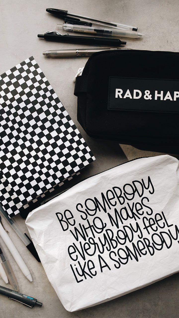 Artistic shot of the white tyvek zipper pouch in the lower part of the photo with the black rad & happy zipper pouch in the upper part sitting on a gray surface surrounded by pens and a sketchbook