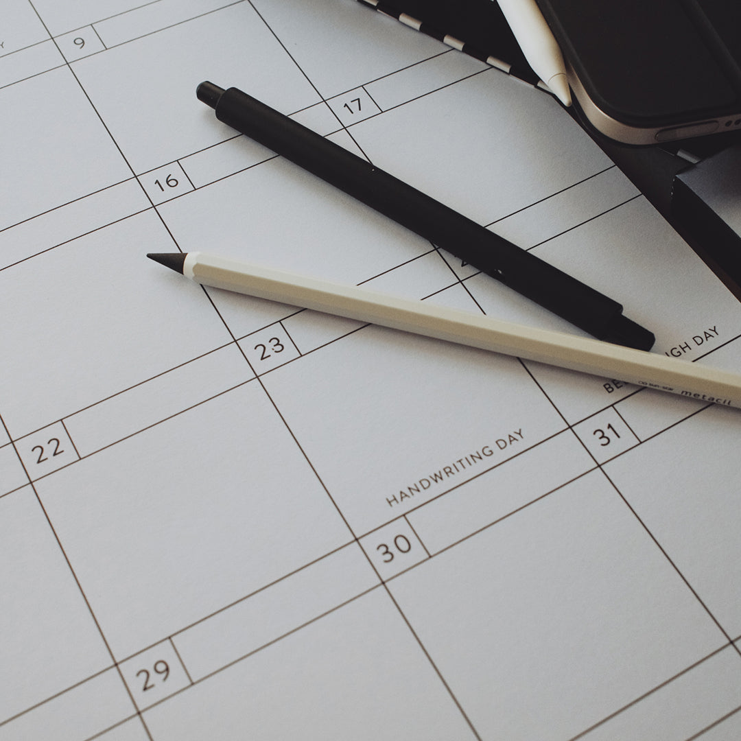 Pens laying on a calendar with the focus on a specific date, showing "Handwriting Day" written on it
