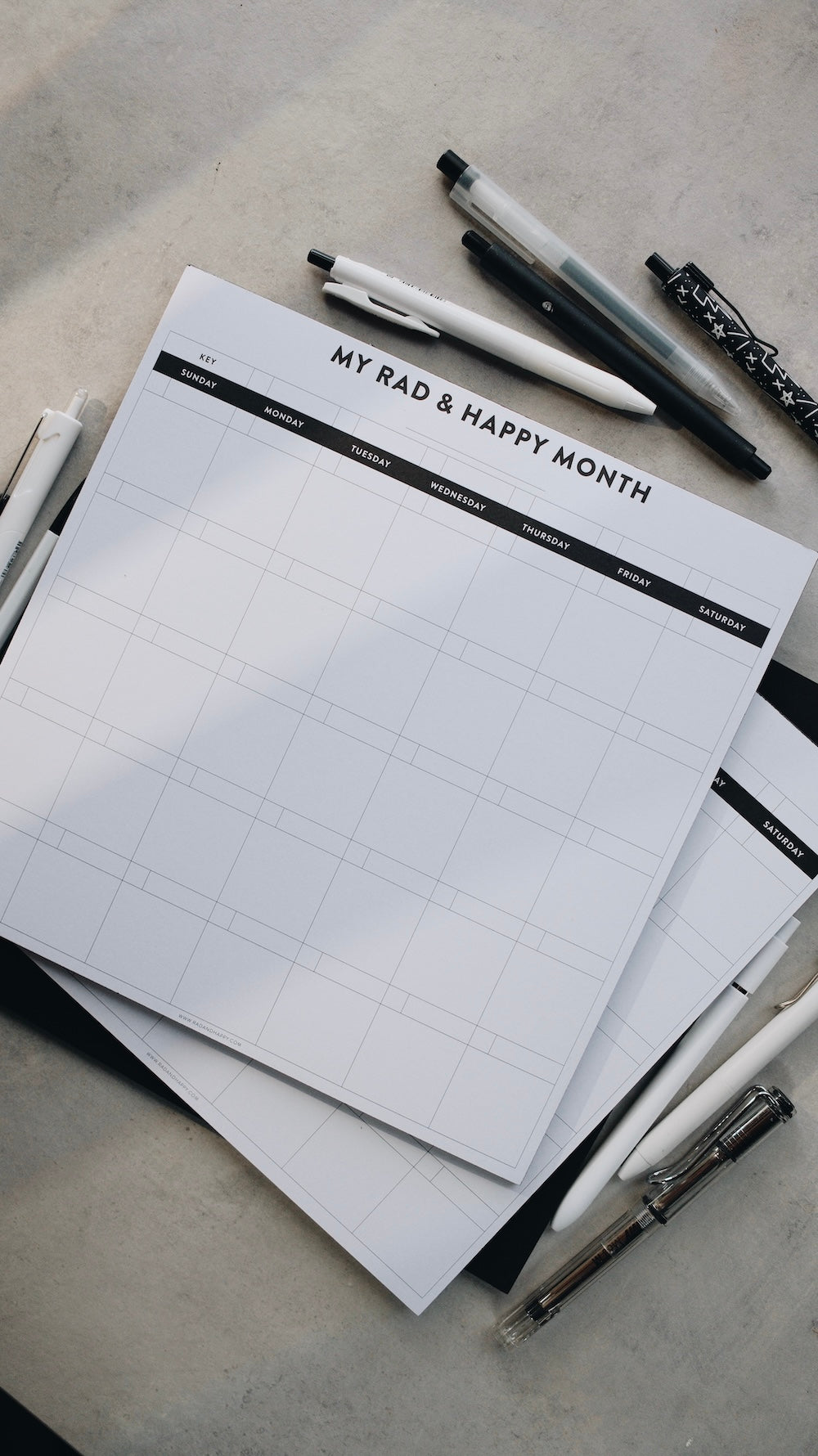 Stack of month notepads with grid layout and text, accompanied by pens on a gray background