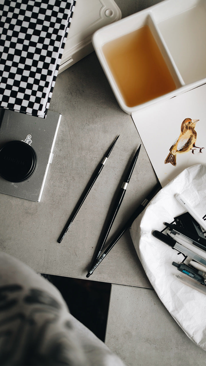 Artistic shot of the corner of the white tyvek zipper pouch with pens on it surrounded by water color tools and a painting of a bird