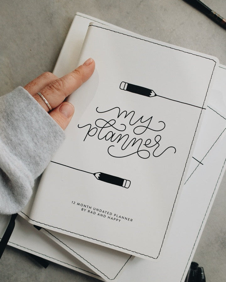 A hand wearing a gray long-sleeve shirt holds an open white planner showing the cover page with black script text reading 'my planner' and pencil illustrations. Other pens and stacked planner pages are visible on a gray tabletop.