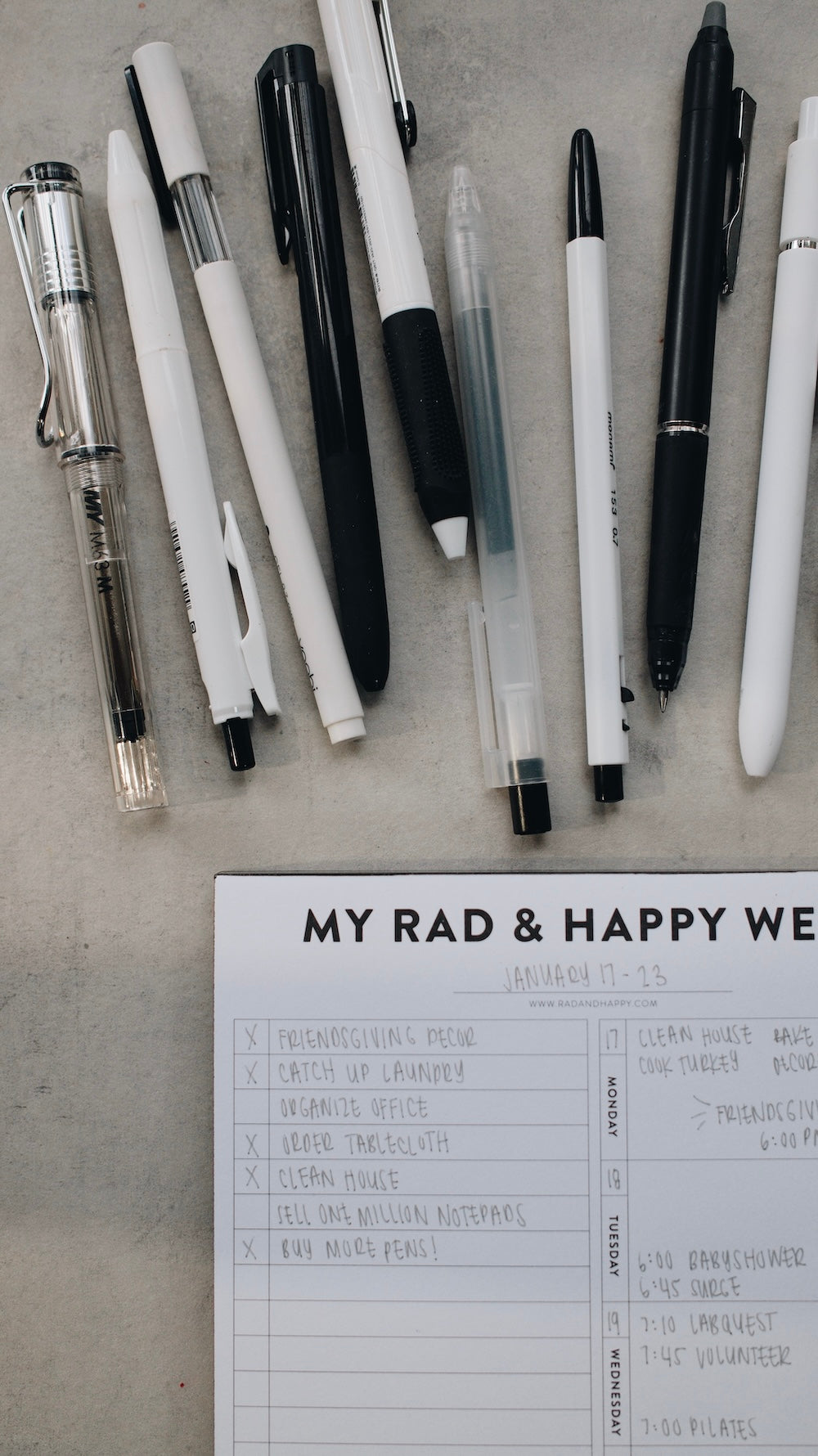 Collection of pens on a concrete surface with a notepad titled 'My Rad & Happy Week'.