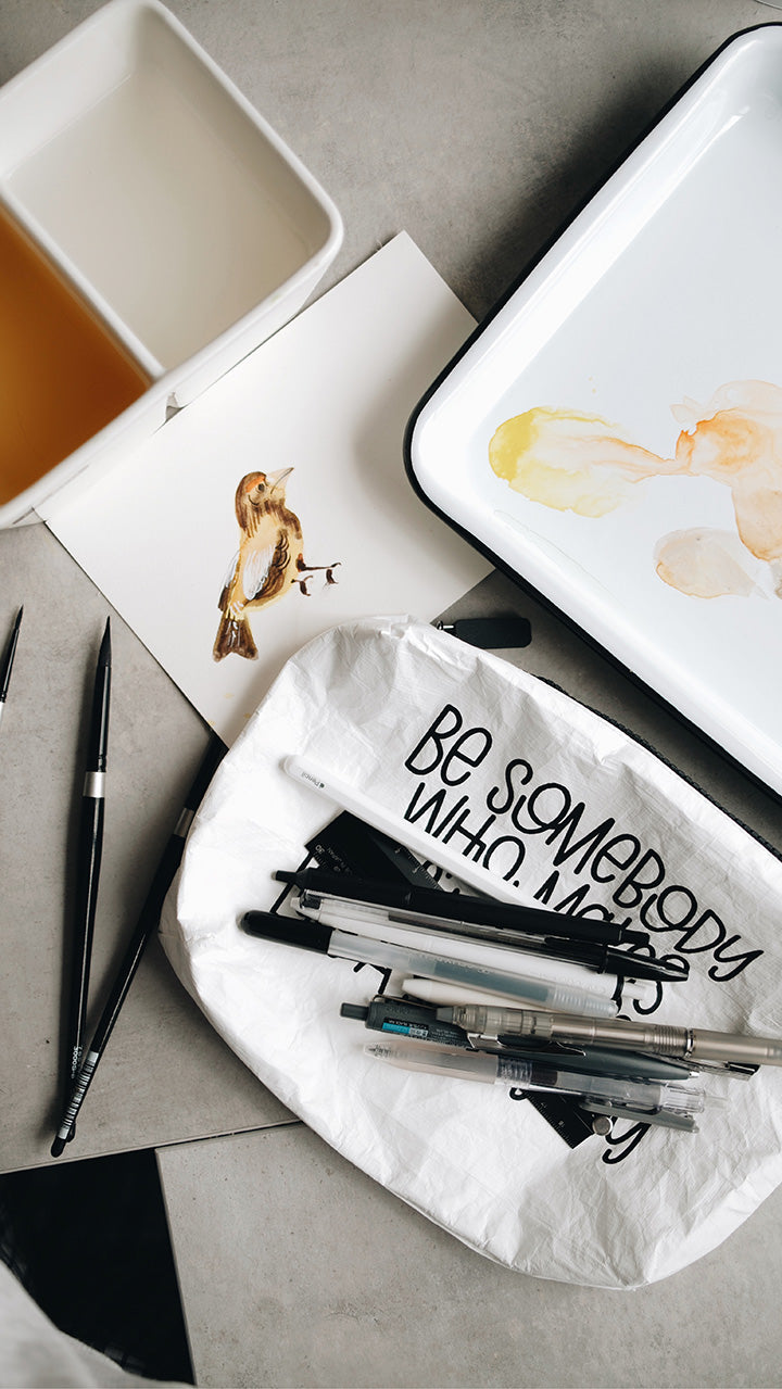 White tyvek zipper pouch with pens laying on it on a gray surface surrounded by water color brushes and a painting of a bird