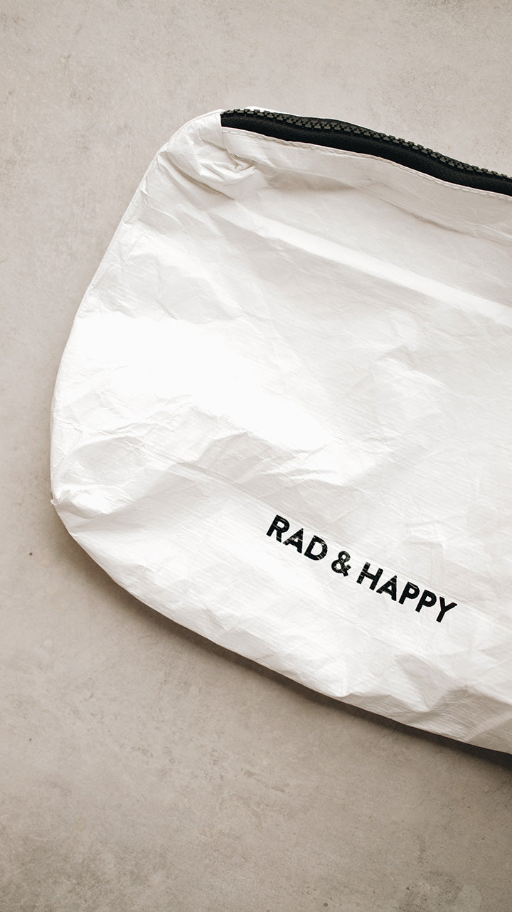 Top corner of a white tyvek zipper pouch with the text Rad & Happy written in black sitting on a gray surface