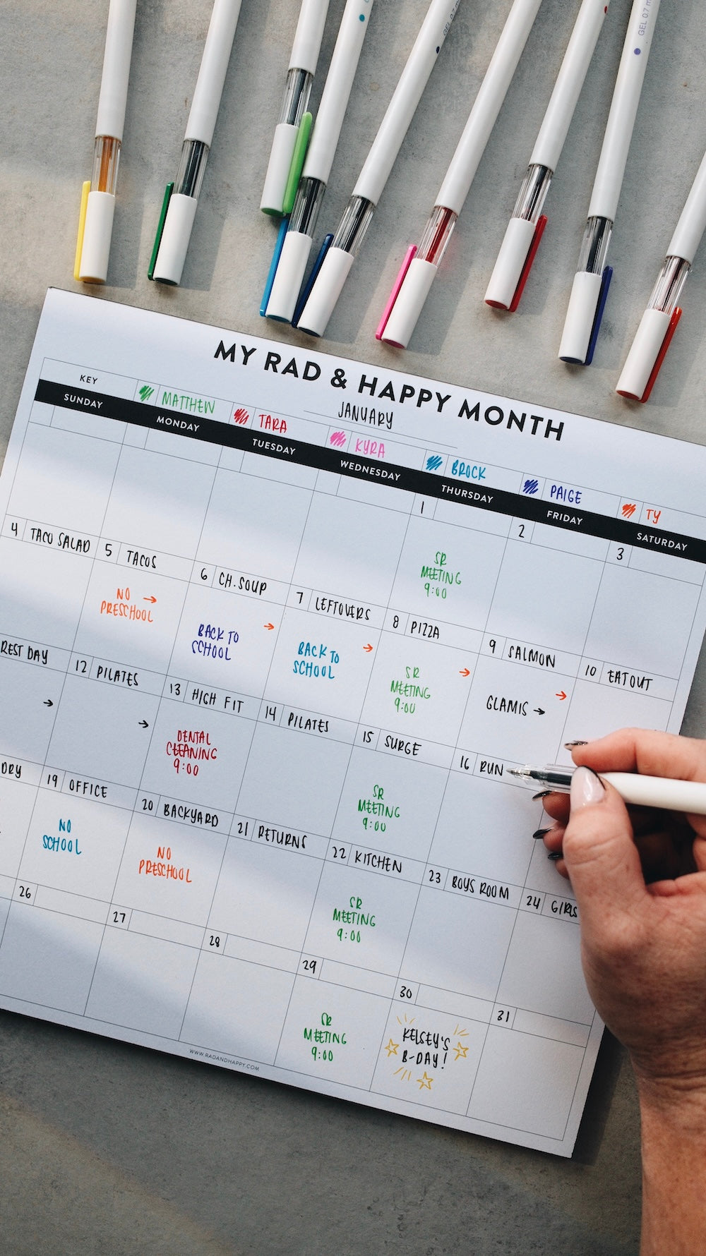 Person writing on a month notepad with markers on a gray surface