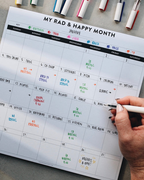 Person writing on a month notepad with markers on a gray surface