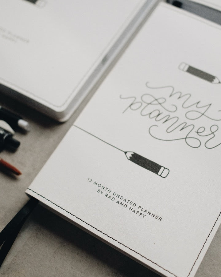 A white leather cover planner with elegant black script reading 'my planner' and a pencil illustration, styled on a gray surface with other notebooks and pens.