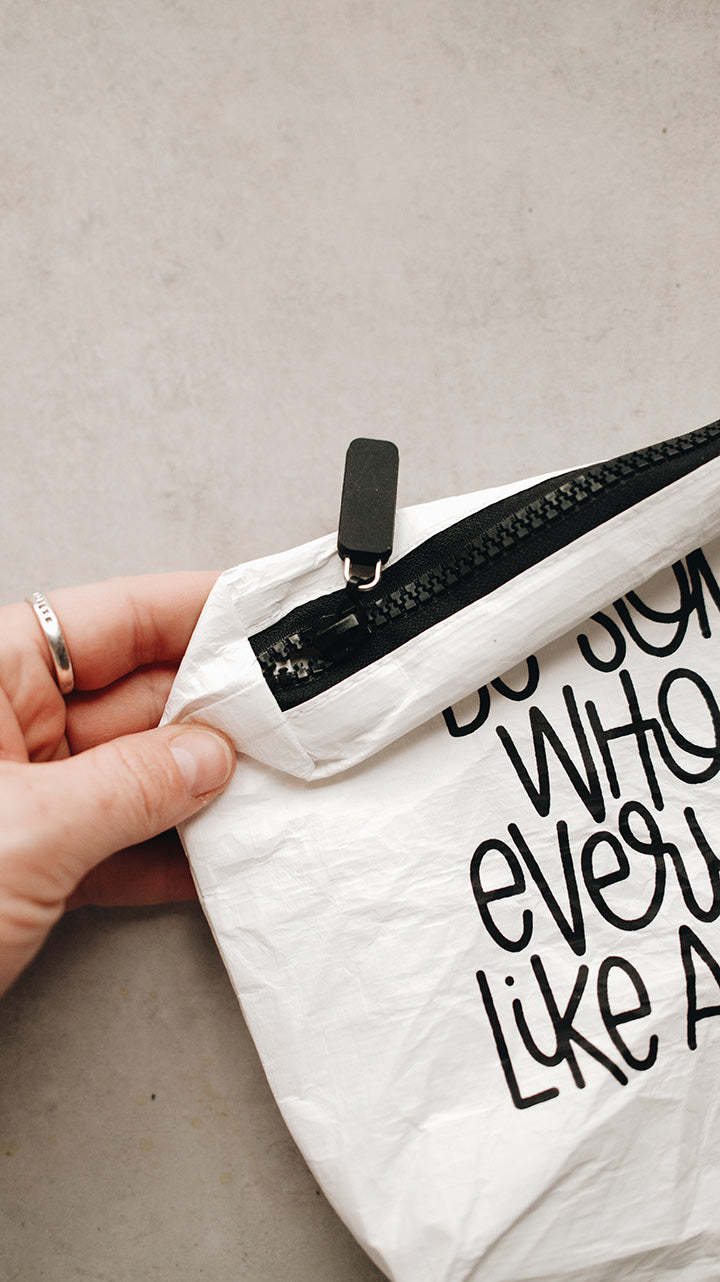 A hand holding the corner of the white tyvek zipper pouch showing the black zipper on a gray surface