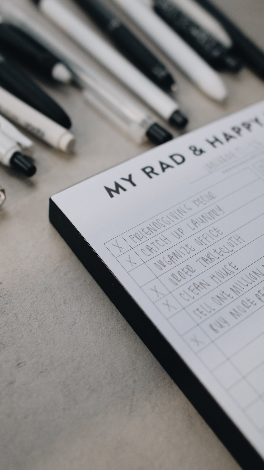 Artistic shot of the corner of a notepad titled 'My Rad & Happy' with a list on a surface with pens.