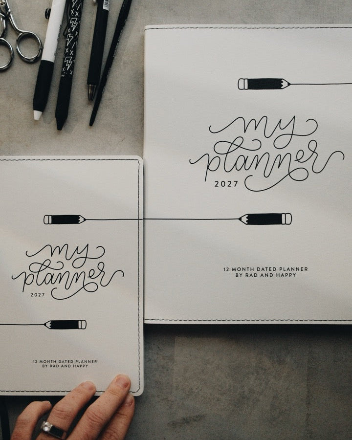 Two white leather-bound planner covers laid on a gray surface, one flat and one held by a hand. The covers feature black script reading 'my planner 2027' with simple line drawings of a pencil and ruler.