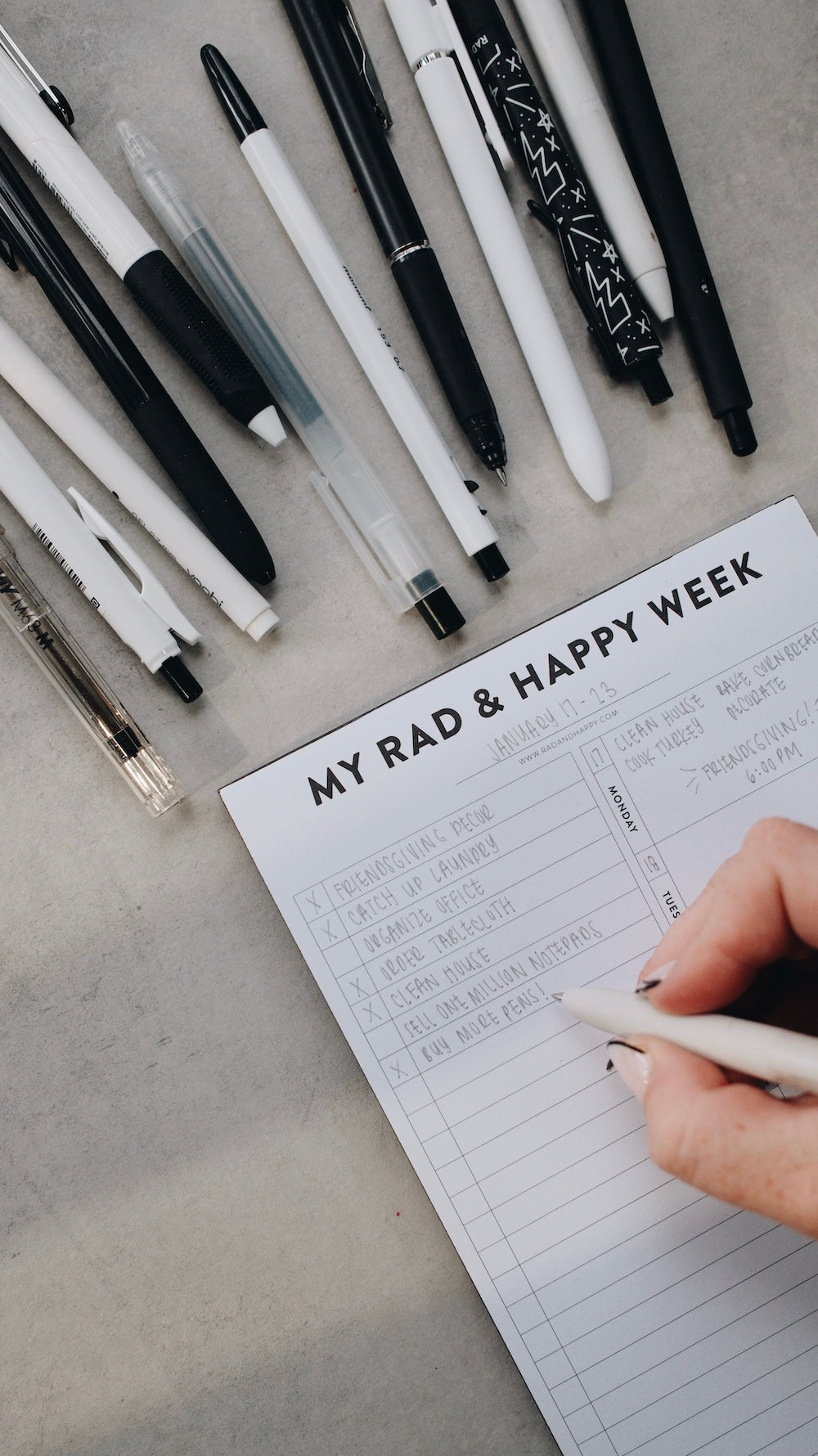 Hand writing on a notebook titled 'My Rad & Happy Week' with pens on a concrete surface