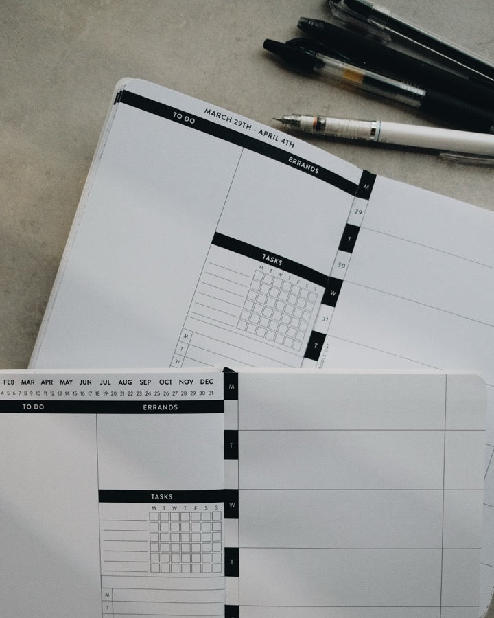 A close-up of a white planner insert with black printed grid lines and tabs, with another matching insert and several pens arranged on a gray surface in the background.