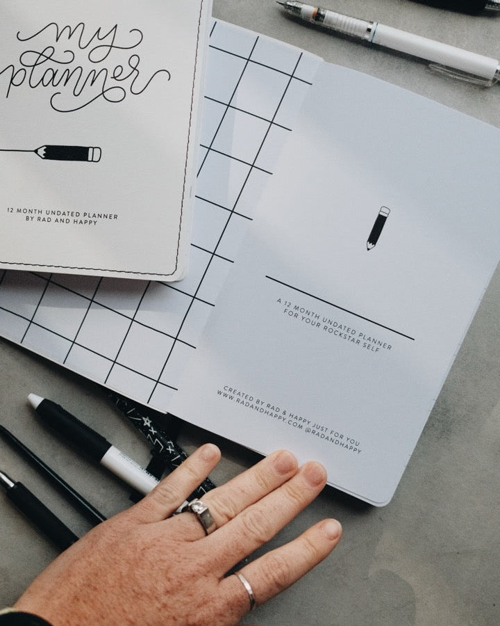 A top-down view of a white monthly planner with 'my planner' written in elegant script, placed on a gray textured surface alongside pens, a black marker, and a person's hand resting near it.