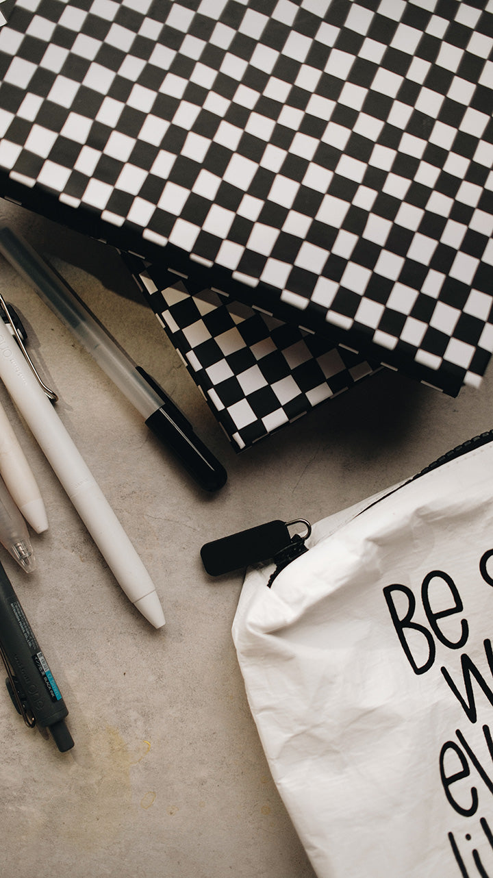 Artistic shot of the corner of the white zipper pouch in the lower part of the photo with a stack of sketchbooks in the upper half of the photo and pens on a gray surface