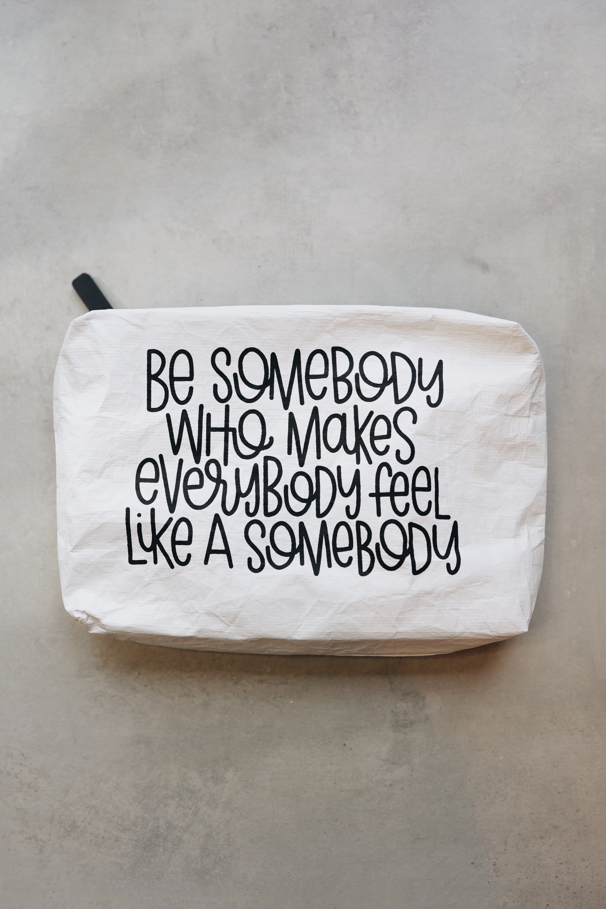 A white Tyvek zipper pouch with the text 'BE SOMEBODY WHO MAKES EVERYBODY FEEL LIKE A SOMEBODY' written in black.