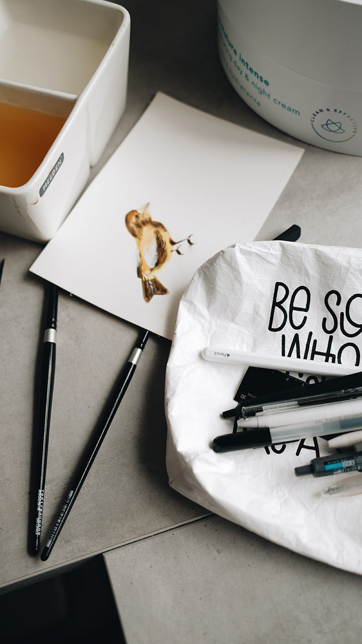 Artistic shot of the left half of the white tyvek zipper pouch with pens laying on it surrounded by water coloring tools and a painting of a bird