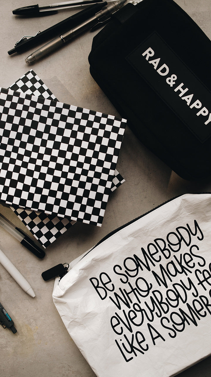 Artistic shot of the white tyvek zipper pouch in the lower part of the photo with the black rad & happy zipper pouch in the upper part sitting on a gray surface surrounded by pens and a sketchbook