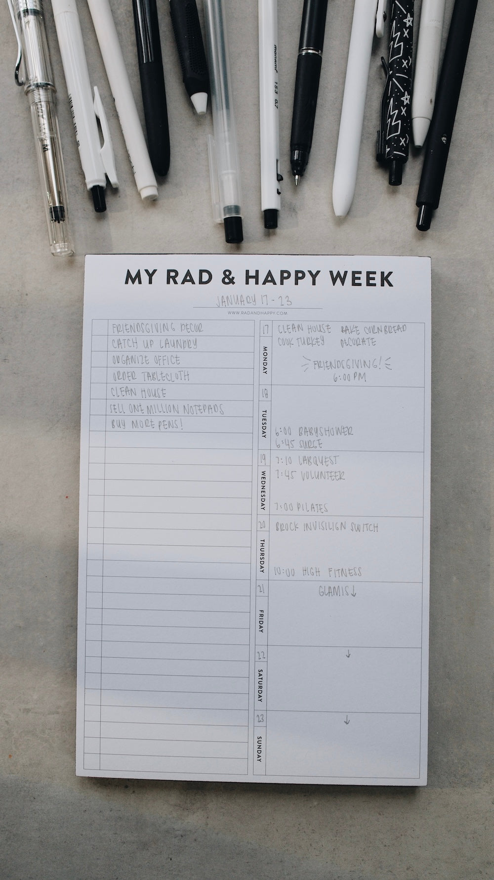 Notepad titled 'My Rad & Happy Week' with pens on a gray surface