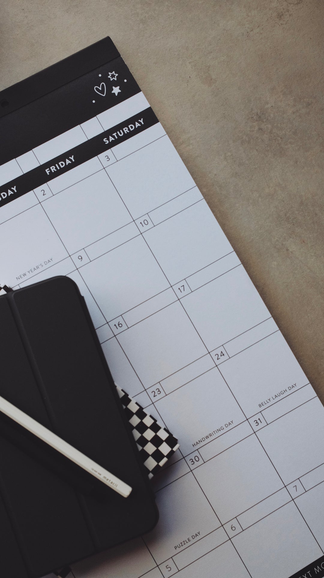 Right-hand corner of a calendar with heart and star doodles lying on a gray surface with a planner and a pen stacked on the calendar