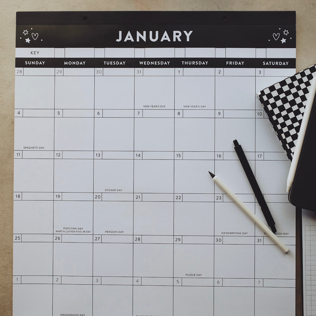 Calendar of the month of January with pens and a planner lying on top of it with a gray background