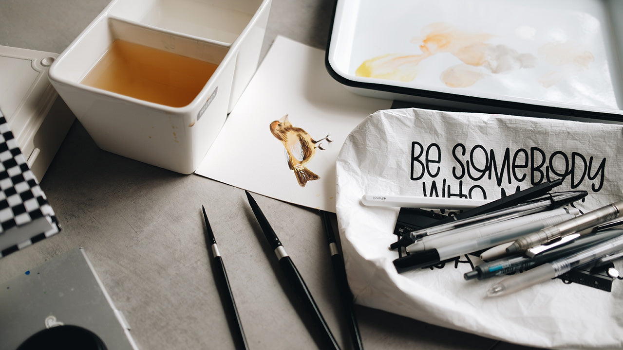 The Rad & Happy white zipper pouch on a desk with pens, pencils, and watercolor brushes around it.