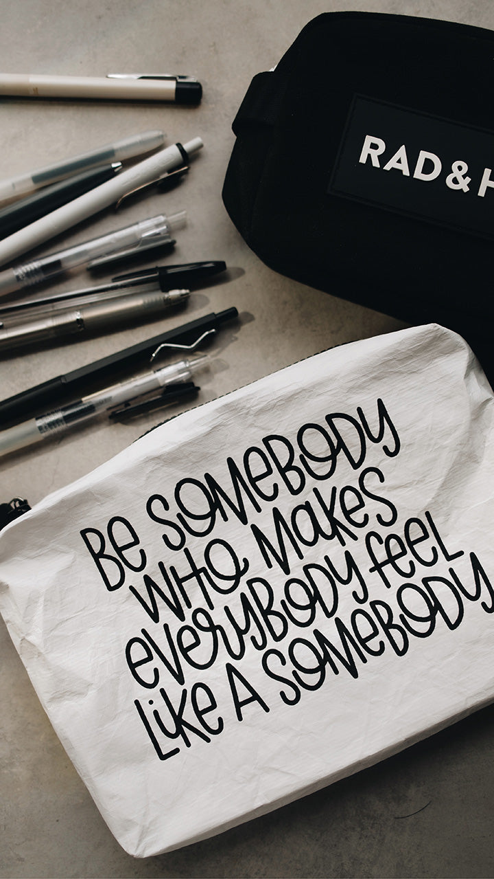 Artistic shot of the white tyvek zipper pouch in the lower part of the photo with the black rad & happy zipper pouch in the upper part sitting on a gray surface surrounded by pens
