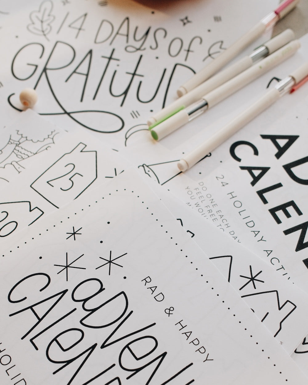 Stack of sheets with '14 Days of Gratitude' and 'Rad & Happy Advent Calendar' text, with markers on a white background.